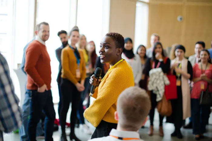 A smiling Black woman with a microphone speaks to a roomful of attendees at a community event