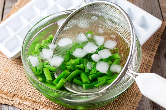 glass bowl of green beans in ice water with strainer scooping the green beans