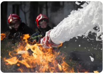flames in the foreground with fire fighters spraying the flames with white foam