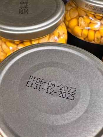 A closeup look at the metal lid of clear jars of corn showing a production date and an expiration date.