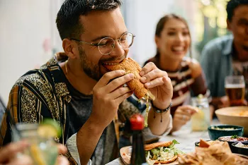 Man taking large bite of a taco with woman smiling in the background