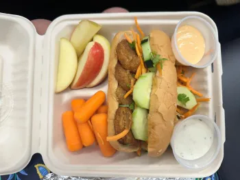 On the menu: Asian Chickenless Veggie subs! Photo shows a to-go box lunch with apples, carrots, and a "chick'n" sub on whole-grain roll with cucumber and shredded carrots, with two dipping sauce options.