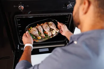 man putting glass dish with chicken legs and vegetables into oven