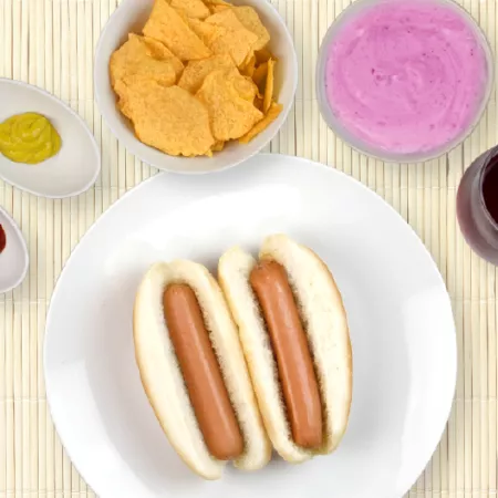 2 hot dogs on a plate surrounded by dips, bowl of chips, blueberry yogurt and cranberry juice