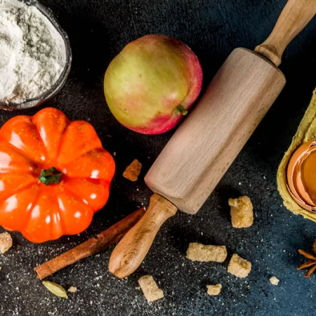 Black background with bowl of flour, small orange pumpkin, apple, rolling pin, carton with one cracked egg, and spices sprinkled in background