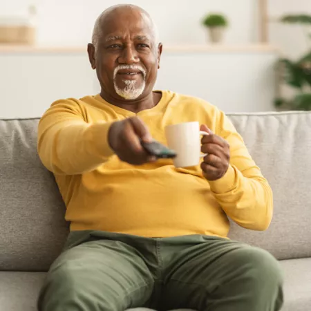 older man holding coffee and pointing remote towards the tv while sitting on a sofa