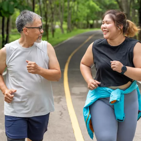 Man and woman with larger bodies unning on a paved trail in a tree lined park