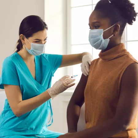 healthcare worker in blue scrubs giving a woman a vaccine shot in her upper arm in front of a bright window