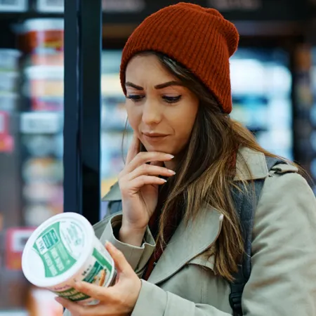 Woman looking confused at back of food container in fridge isle of the grocery store