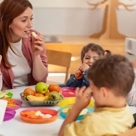 A female teacher sits with young students at a classroom table sharing snacks free from the Big Nine allergens. 